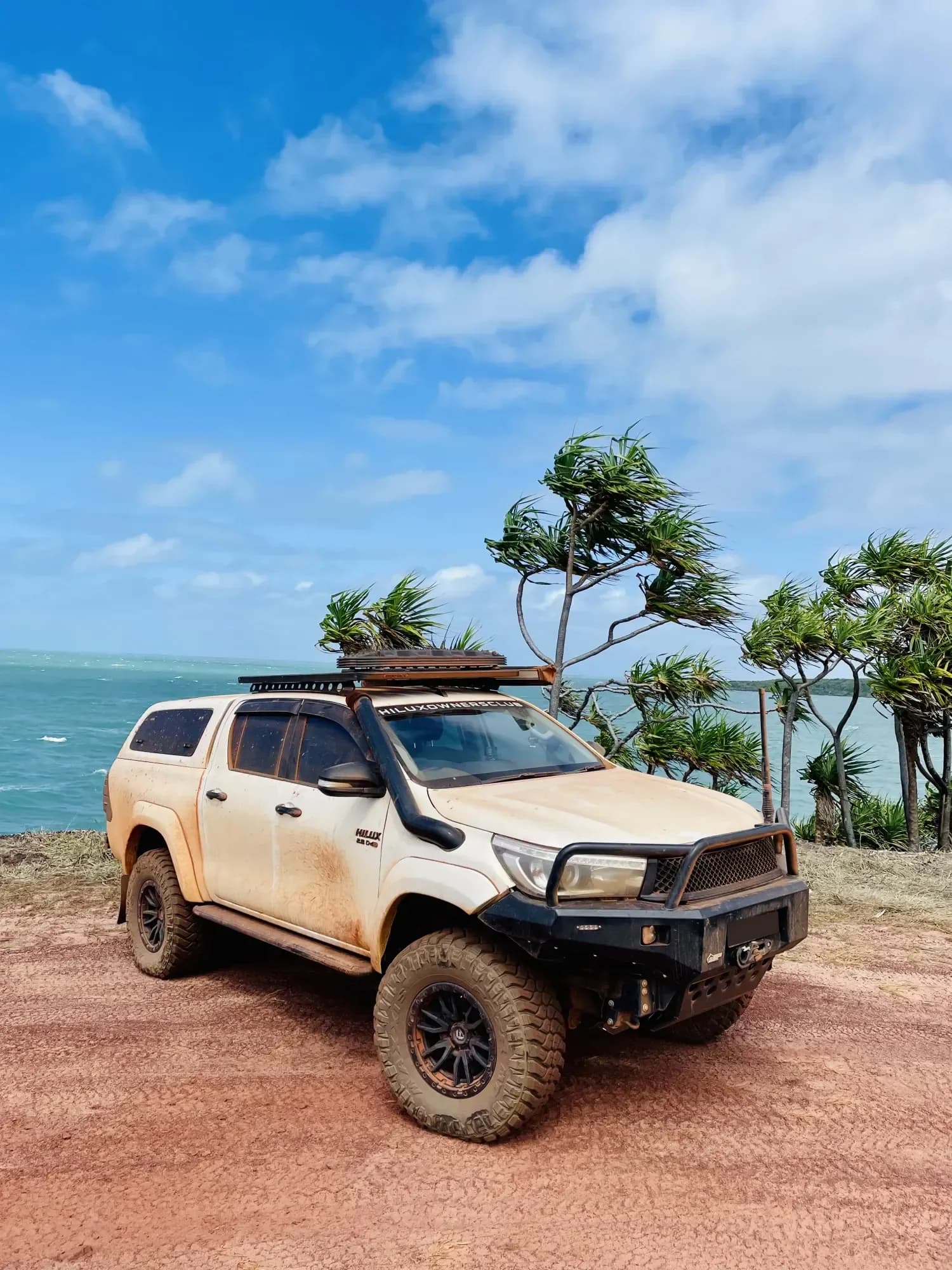 A dusty white Hilux with a roof rack parked on a clifftop beside pandanus palms above a turquoise sea